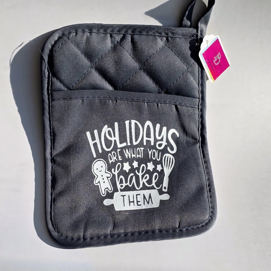 A dark gray quilted oven mitt with a front pocket featuring the white printed text 'HOLIDAYS ARE WHAT YOU bake THEM' along with a gingerbread man and whisk illustration. A small pink tag is attached near the top edge, and the item is photographed on a light gray surface with a hard shadow.