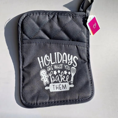 A dark gray quilted oven mitt with a front pocket featuring the white printed text 'HOLIDAYS ARE WHAT YOU bake THEM' along with a gingerbread man and whisk illustration. A small pink tag is attached near the top edge, and the item is photographed on a light gray surface with a hard shadow.