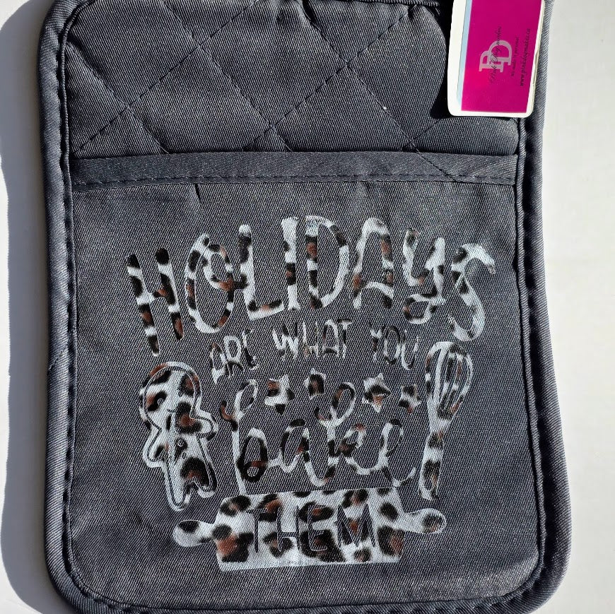 A dark gray quilted oven mitt featuring a leopard-print patterned graphic that reads 'HOLIDAYS ARE WHAT YOU BAKE THEM' with baking utensil illustrations. A pink and white tag is attached to the top right corner.