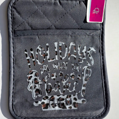 A dark gray quilted oven mitt featuring a leopard-print patterned graphic that reads 'HOLIDAYS ARE WHAT YOU BAKE THEM' with baking utensil illustrations. A pink and white tag is attached to the top right corner.