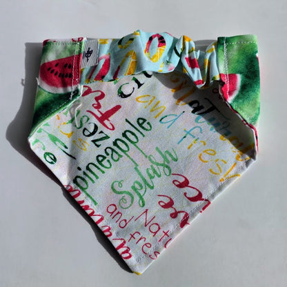A fabric pet bandana with a colorful watermelon and citrus print on one side and a white background with multicolored script words like 'pineapple', 'ice', and 'fresh' on the other, shown on a plain light background.