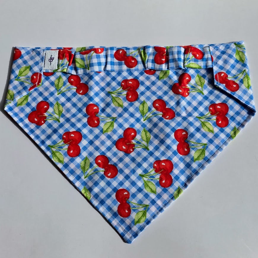 A triangular pet bandana with a blue and white gingham pattern overlaid with a red cherry and green leaf print, laid flat on a light gray background.