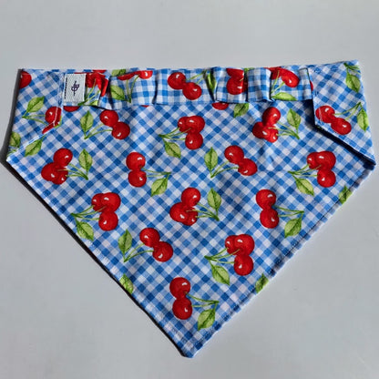 A triangular pet bandana with a blue and white gingham pattern overlaid with a red cherry and green leaf print, laid flat on a light gray background.