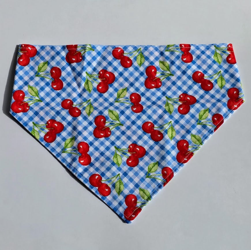 A scrunchie style pet bandana featuring a blue and white gingham check pattern with a repeating print of red cherries and green leaves, laid flat on a light gray background.