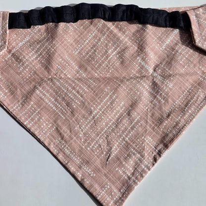 A triangular bandana-style scarf or pet bandana in dusty pink fabric with a white dotted geometric pattern and a black ruffled trim along the top edge, laid flat on a light gray surface.