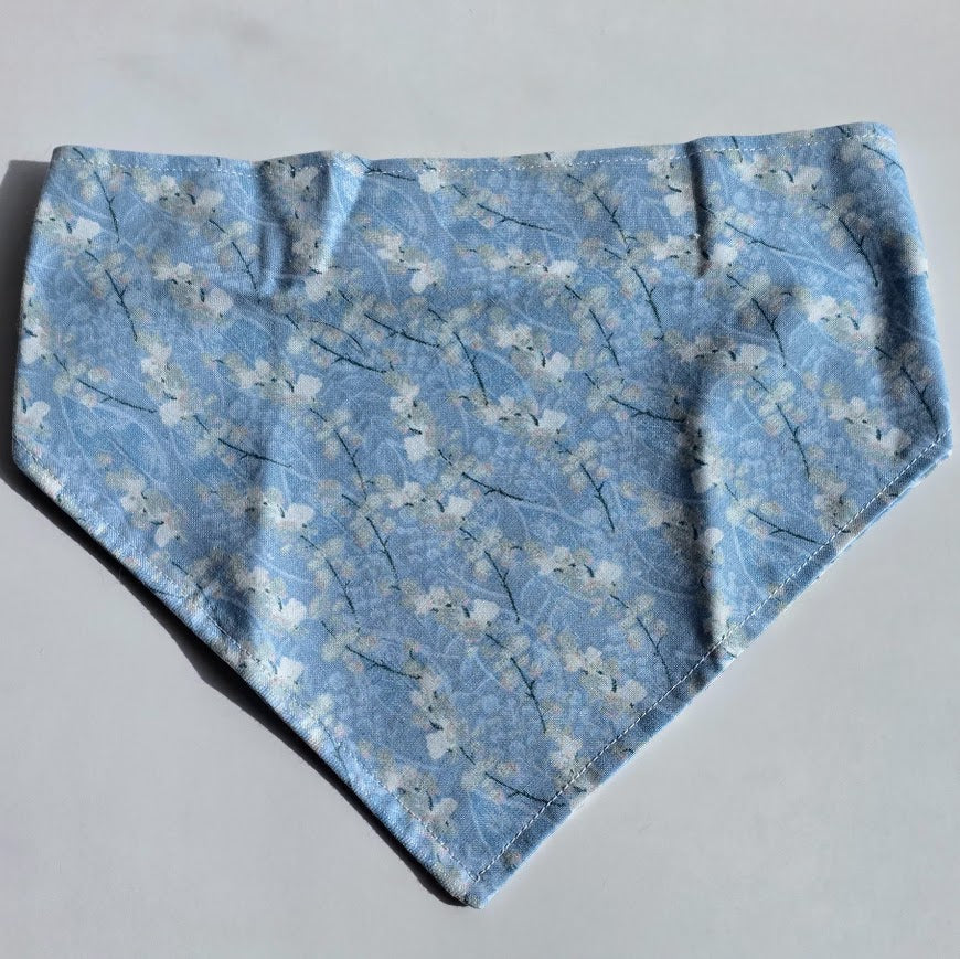 A scrunchie style pet bandana featuring a light blue and white cherry blossom pattern, laid flat on a neutral light gray background.