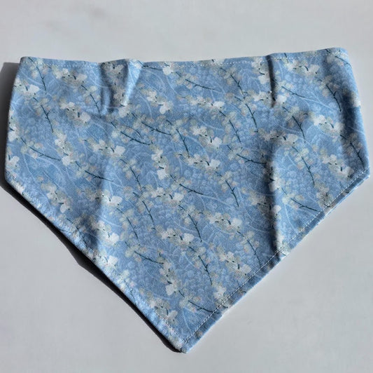 A scrunchie style pet bandana featuring a light blue and white cherry blossom pattern, laid flat on a neutral light gray background.