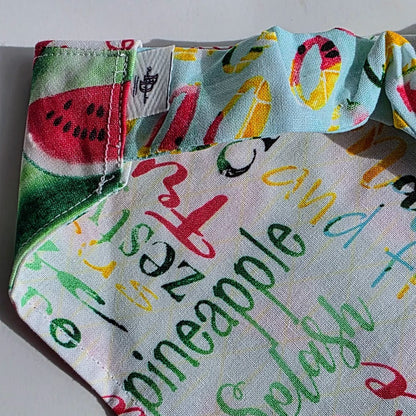 A close-up of a fabric product, likely a swim cap or headband, featuring a colorful summer-themed print with watermelon slices, tropical letters, and words like pineapple, melon, and citrus on a white background.