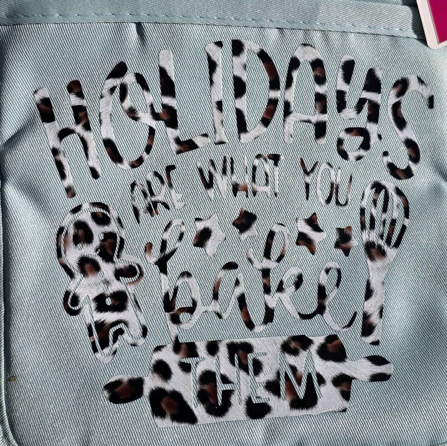 A close-up of a light blue fabric item, likely a tote bag or backpack, featuring a decorative graphic with the text 'HOLIDAYS ARE WHAT YOU BAKE THEM' in a leopard print pattern.