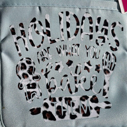 A close-up of a light blue fabric item, likely a tote bag or backpack, featuring a decorative graphic with the text 'HOLIDAYS ARE WHAT YOU BAKE THEM' in a leopard print pattern.