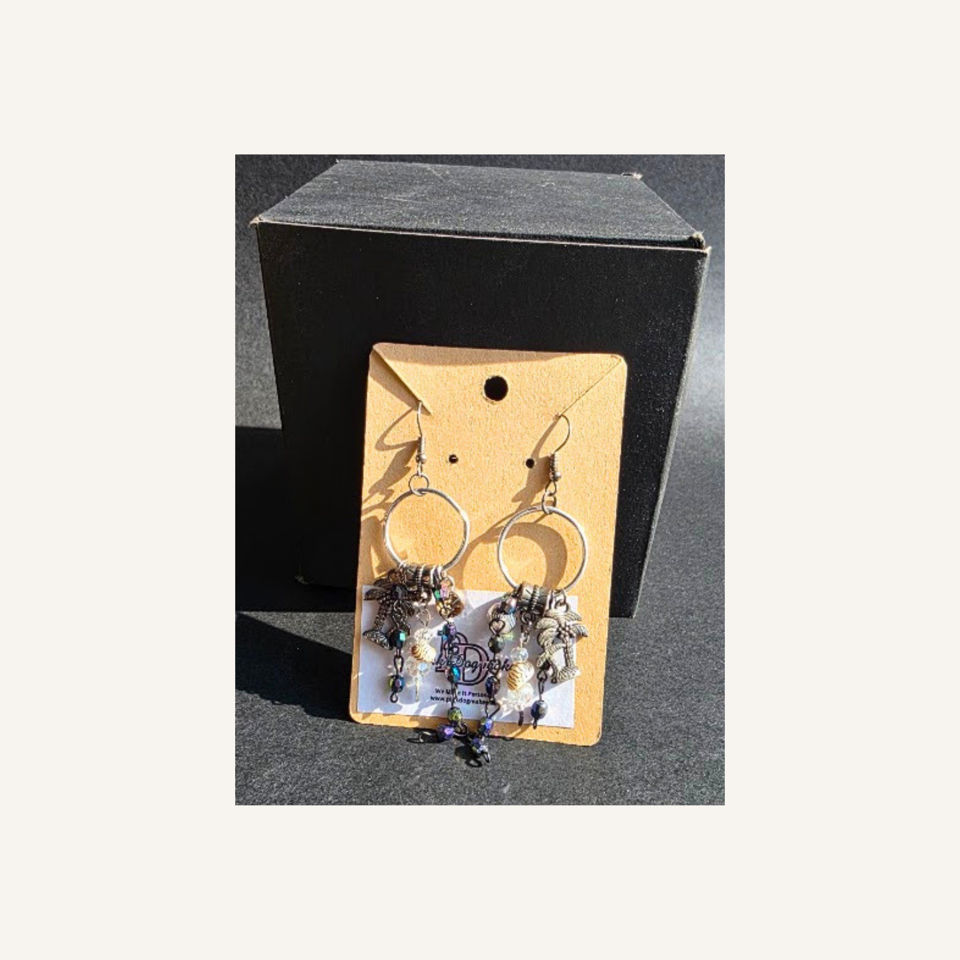 A pair of dangling hoop earrings with layered beaded and metallic details, displayed on a tan card in front of a black box against a dark surface.