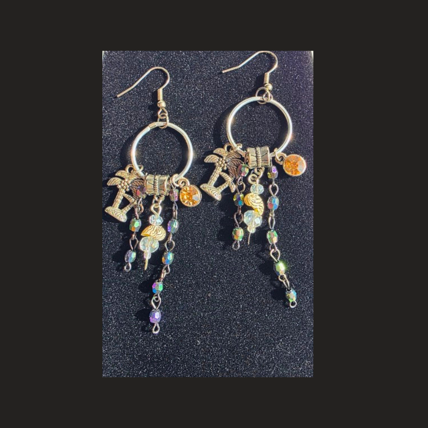 A pair of silver-tone dangle earrings with circular hoops, palm tree charms, a small gold-toned cross charm, and iridescent beads, displayed on a textured black background. Handmade silver-plated tropical charm hoop earrings with colourful dangling beads and palm tree charms