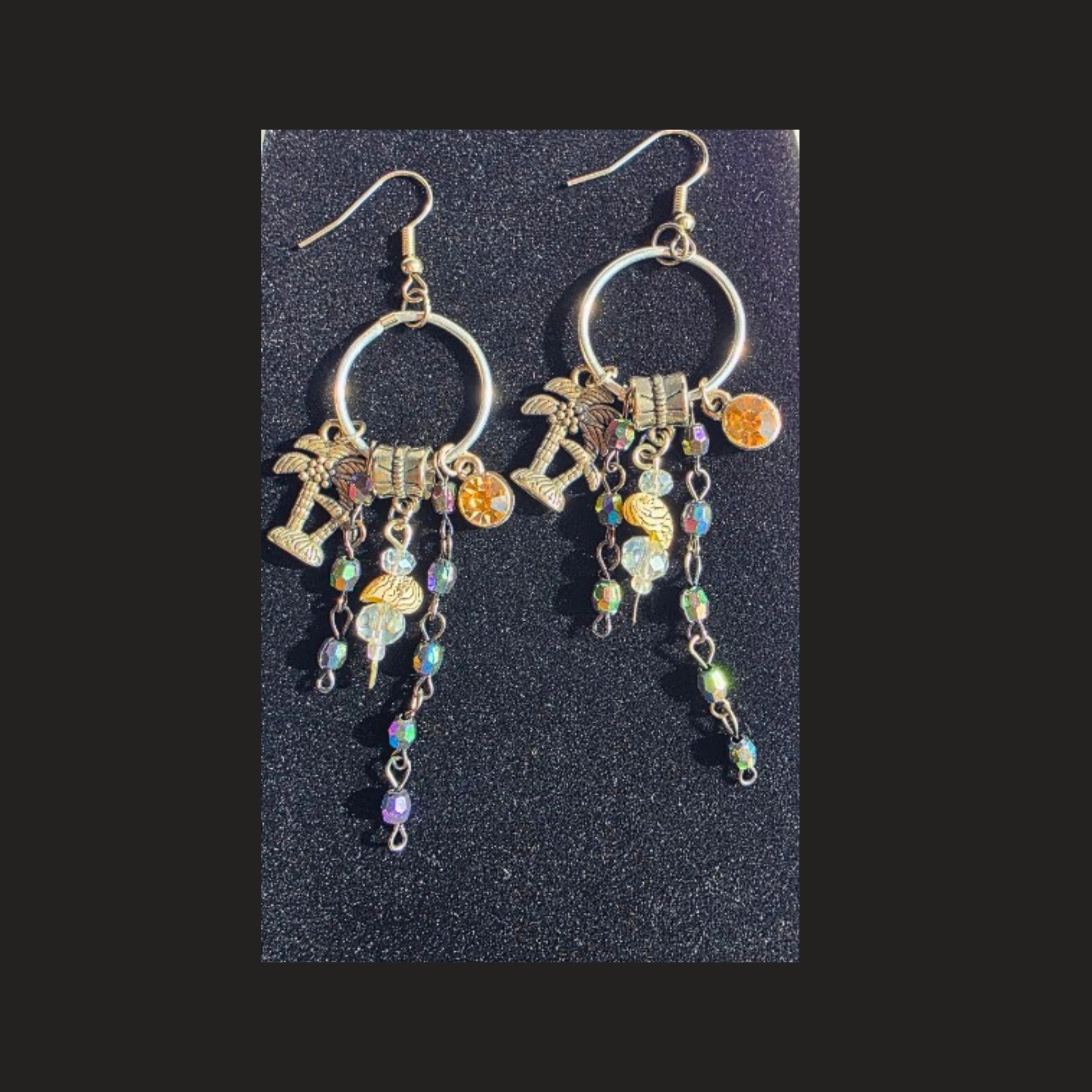 A pair of silver-tone dangle earrings with circular hoops, palm tree charms, a small gold-toned cross charm, and iridescent beads, displayed on a textured black background. Handmade silver-plated tropical charm hoop earrings with colourful dangling beads and palm tree charms