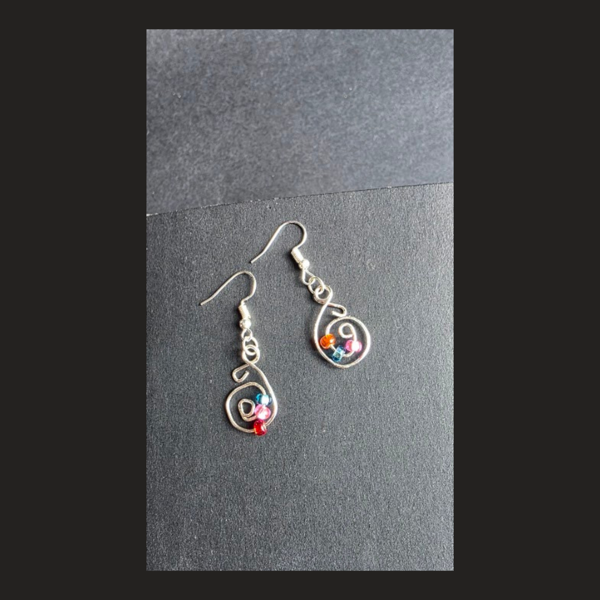 A pair of handmade silver dangle earrings with colorful gemstone accents, displayed on a dark gray textured card against a black background.