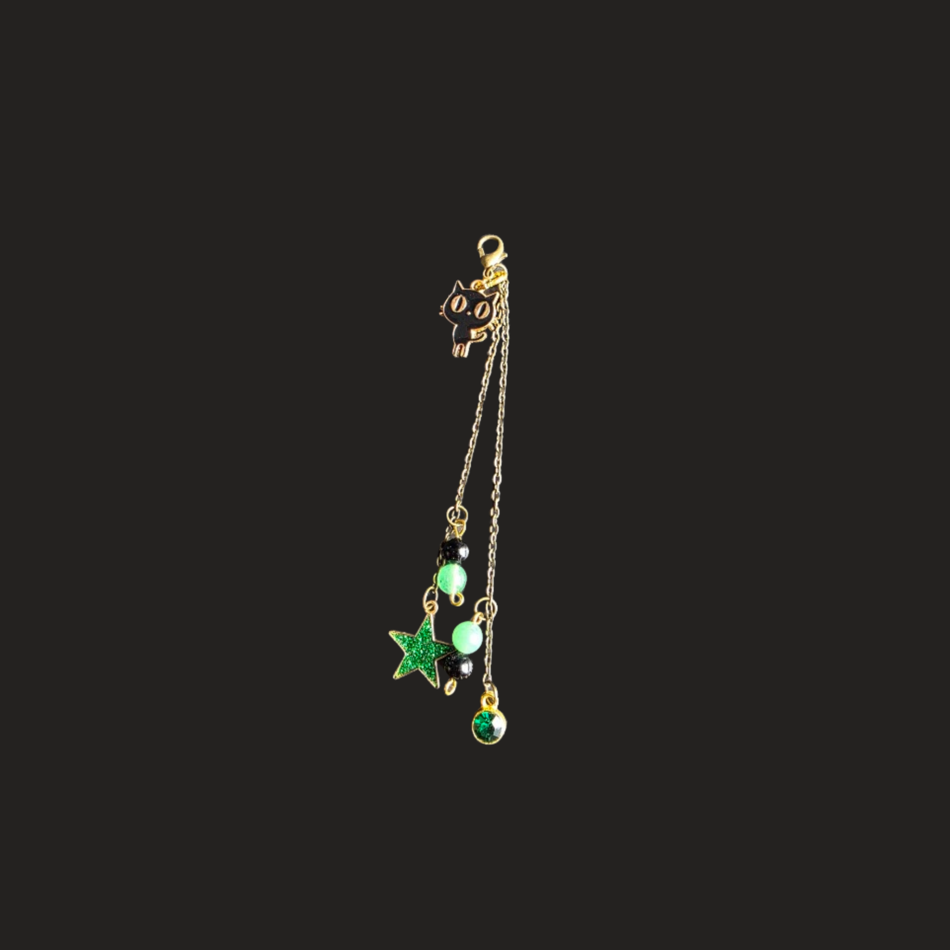 A gold-toned dangle earring featuring a small cat charm, a green star charm, light green beads, and a small round green gem, shown against a solid black background.
