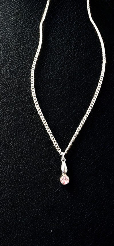 A silver chain necklace with a round-shaped silver pendant holding a small round pink gemstone, displayed on a textured black background.
