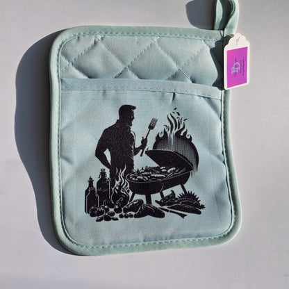 A light blue quilted pot holder with a black graphic of a man grilling, featuring a front pocket and a hanging purple brand tag.