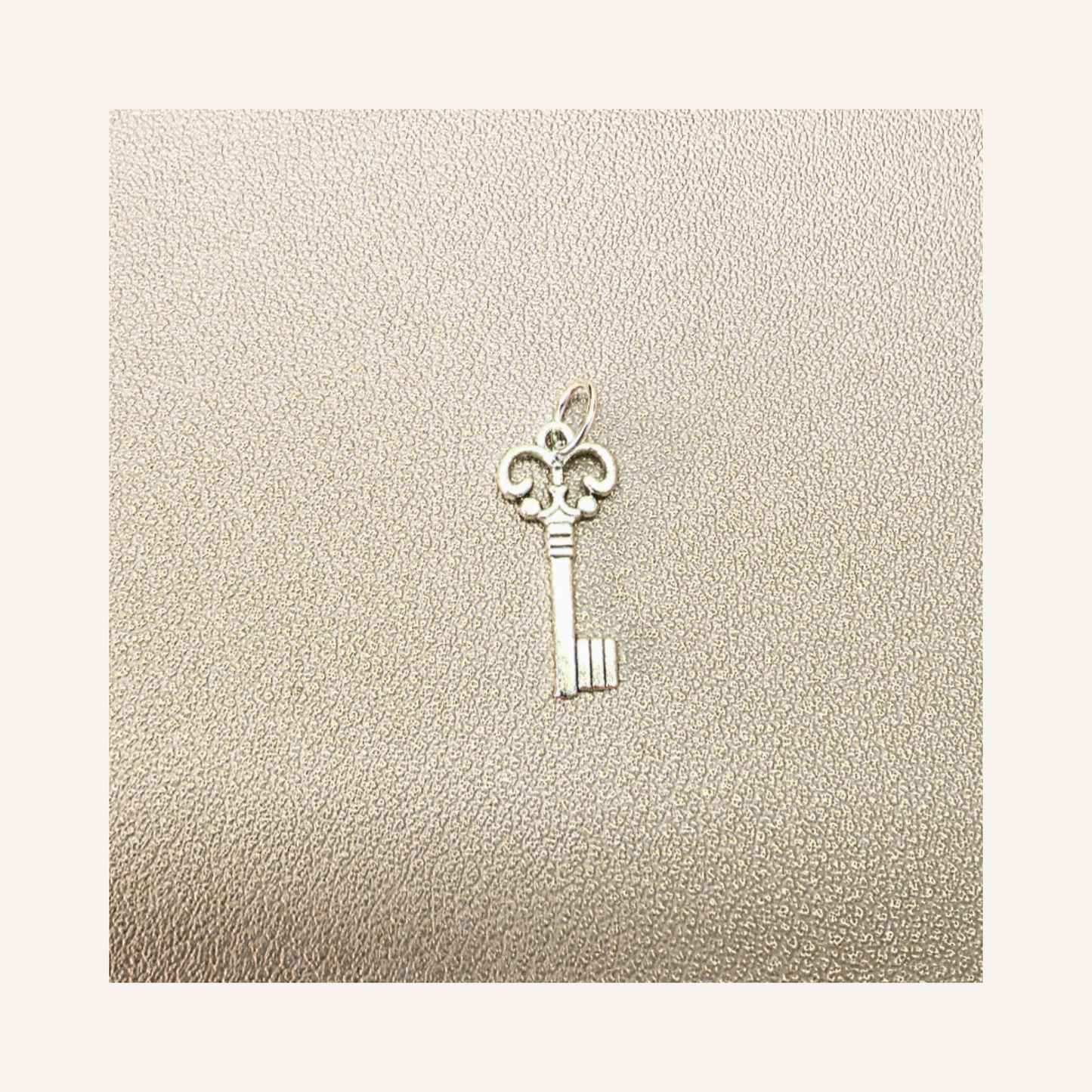A small silver key-shaped pendant or charm displayed on a textured beige surface with a light cream border.