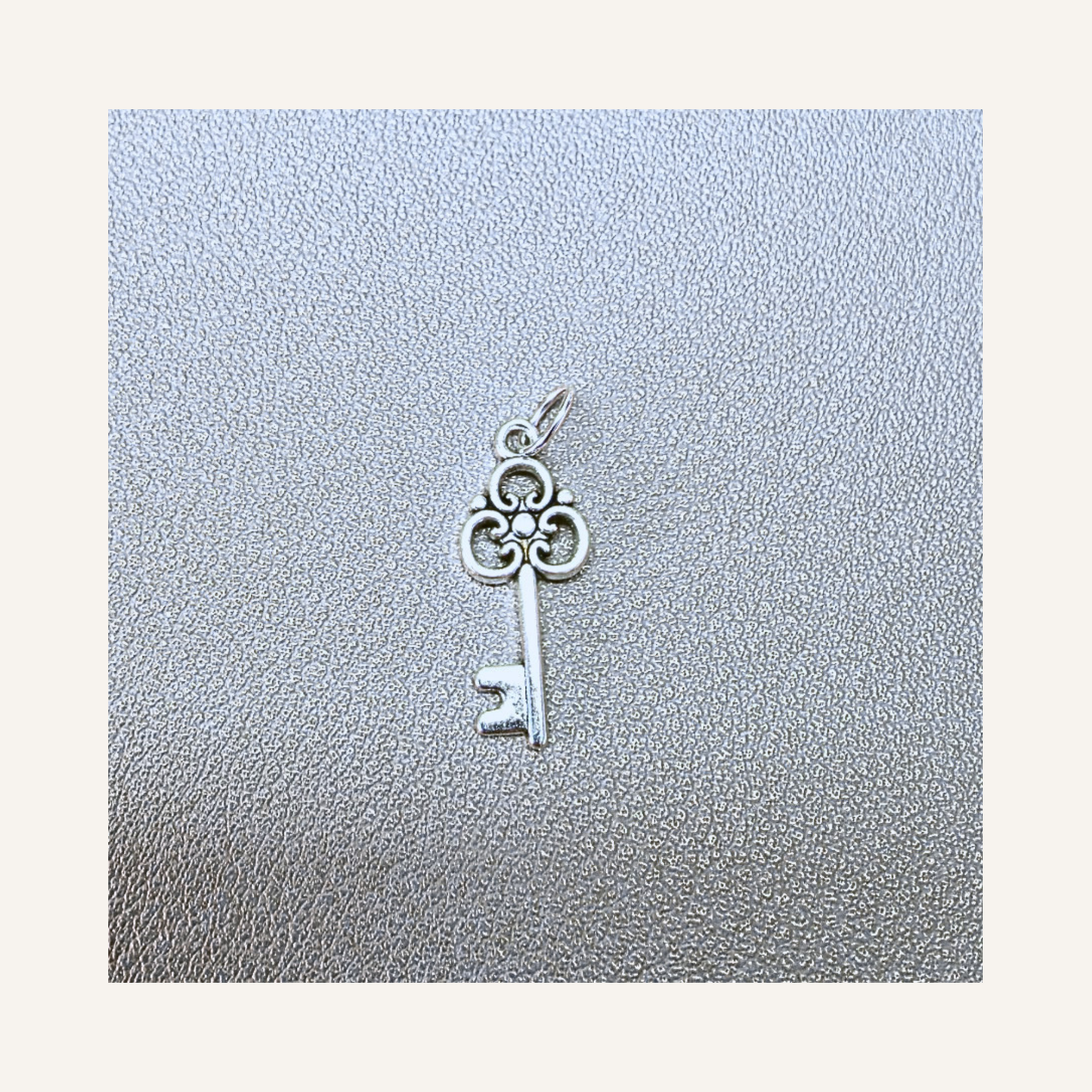 A silver key-shaped charm pendant with a decorative three-loop top and a small bail, displayed on a textured silver surface against a cream background.
