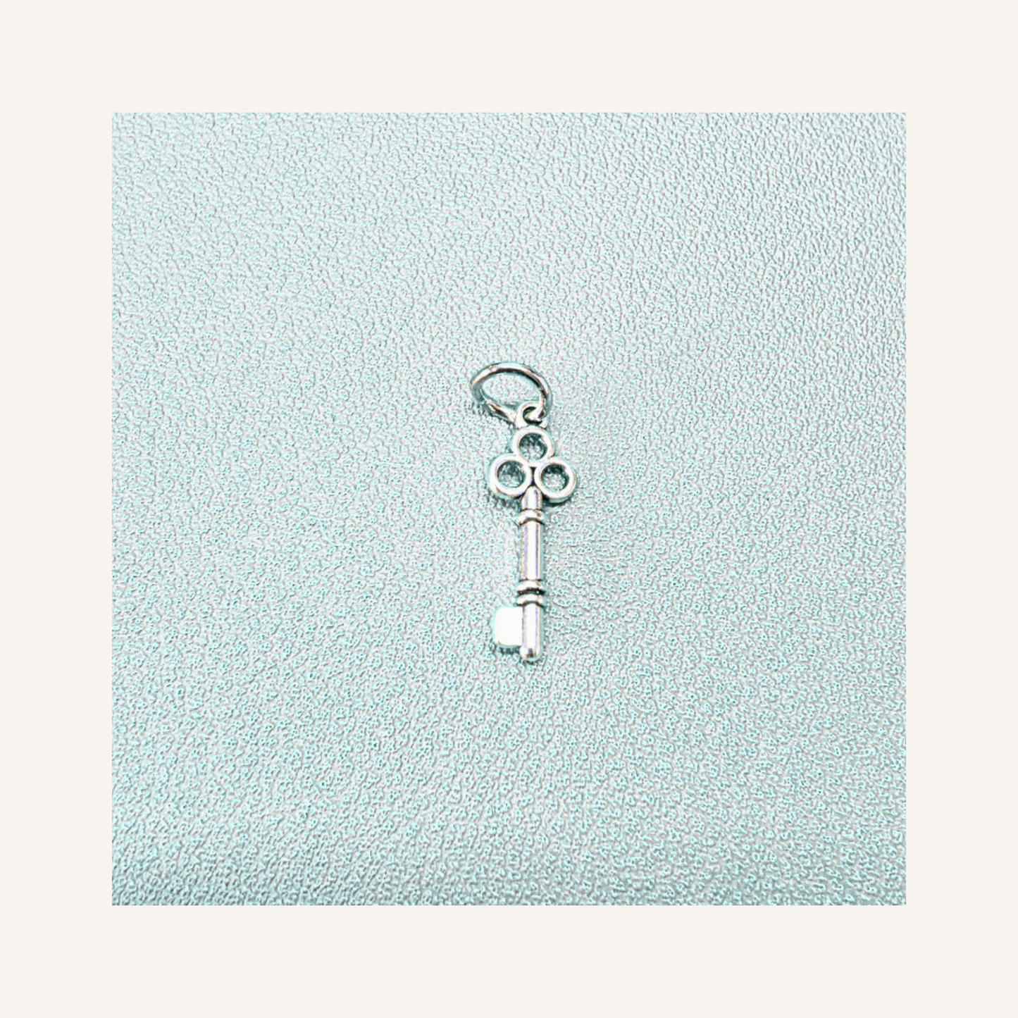 A small silver key-shaped charm with a decorative ring at the top, displayed on a textured light blue square background with a cream border.