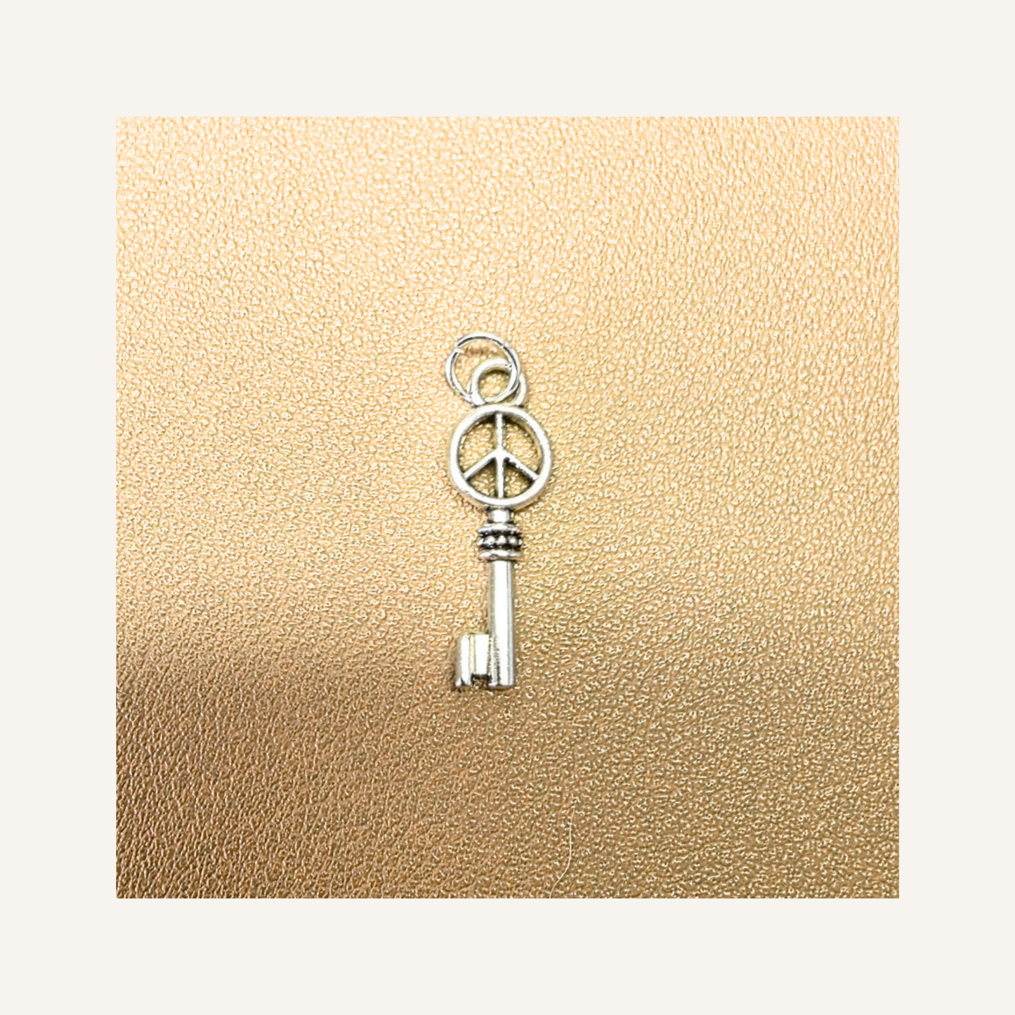 Silver key charm for bracelet or necklace with simple bail and peace symbol at top. Displayed on cream leather back ground.  from pink dog makes mix and match collection.