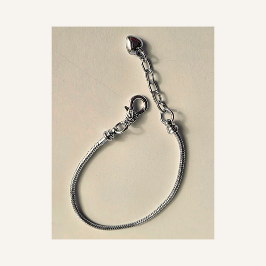 A silver snake chain bracelet with a removeable lobster clasp and an adjustable extension chain featuring a small heart-shaped charm, displayed on a light beige background.