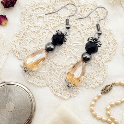 A pair of handmade dangle earrings with silver fishhook wires, black beads, and amber-colored teardrop crystals, styled on a white lace doily with vintage props including dried roses, a pearl necklace, and a handkerchief.