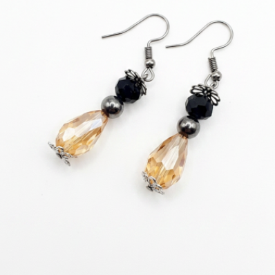 A pair of dangle earrings with silver fishhook wires, small black beads, dark gray metallic beads, and faceted amber-orange teardrop crystal drops, shown on a plain white background.