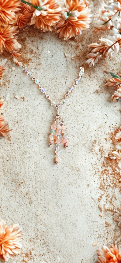 A delicate necklace with a multi-colored pastel pendant featuring pink, orange, and blue hues, styled on a textured beige surface surrounded by dried orange flowers.