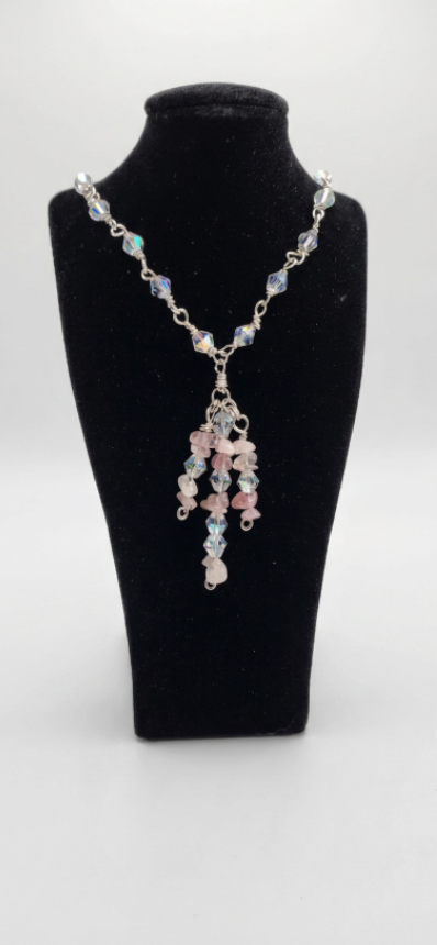 A beaded necklace displayed on a black velvet jewelry bust against a light gray background. The necklace features silver-toned chain links and beads in pastel pink, light blue, and iridescent tones, with a dangling cluster of pink and blue beads at the center.