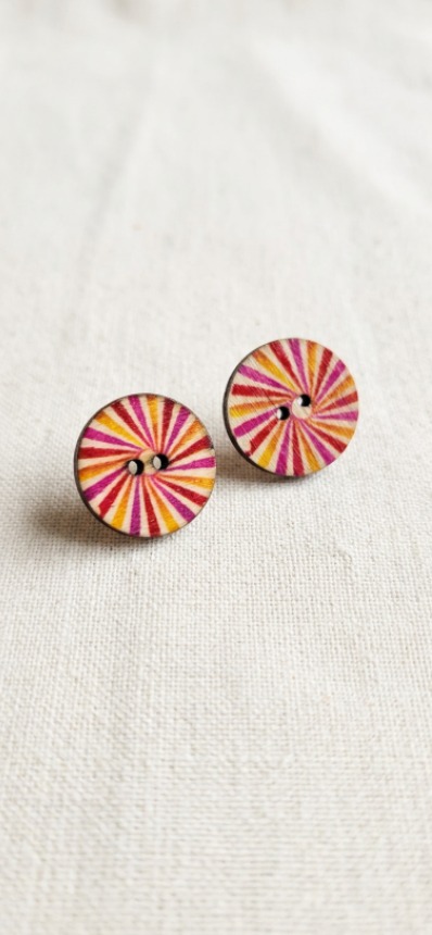 A pair of round wooden buttons with a colorful radial sunburst pattern in pink, orange, yellow, and white, resting on a textured off-white linen surface.